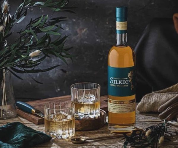 A bottle of whiskey on a table accompanied by two filled glasses on the rocks set in a rustic background with some greenery.