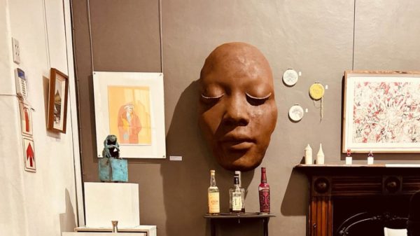 Image of large female face sculpture with framed art on either side and bottles of whisky on a show table infront of the sculpture haging 